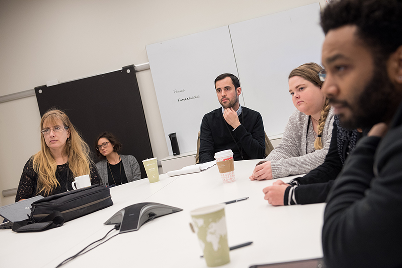 A group of people are seated around a conference table in a meeting room. They appear focused and serious, as if listening or discussing an important topic. Coffee cups, notebooks, and a speakerphone on the table suggest a professional work meeting.