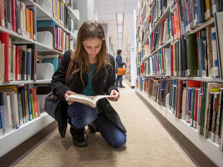 A student kneeling in an aisle at the Iwasaki Library, reading through a book