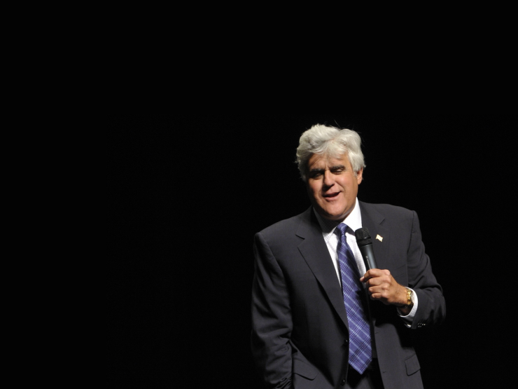 Comedian Jay Leno holding a microphone, standing in front of a black background