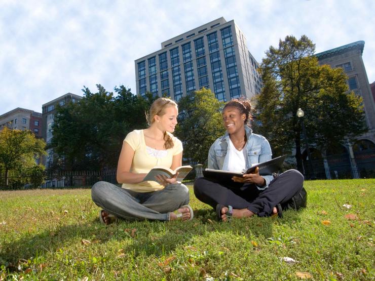 Two students sitting on a lawn with a building behind them