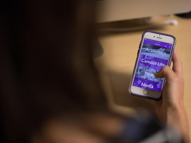 A student holding an iPhone displaying the Emerson College mobile app.