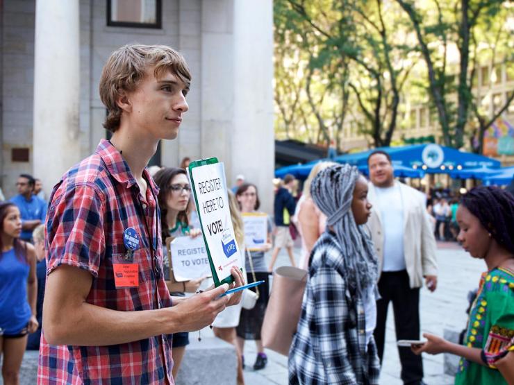 Young person at Faneuil Hall holding up a clipboard reading "Register Here to Vote"