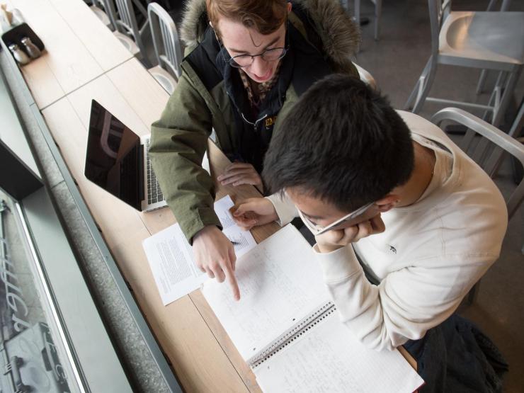 Two students looking over a notebook with one pointing at a page. They are sitting in front of a laptop.