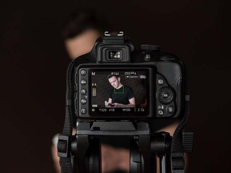 A photo of a young man obscured by a camera in the foreground. The young man can be seen from the camera's digital viewfinder.