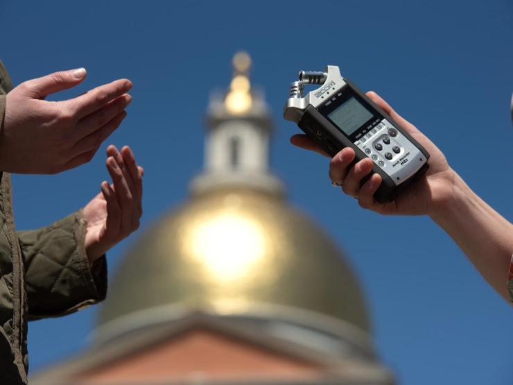 Students conduct an interview in front of the Massachusetts State House. The golden dome of the State House can be seen in the background, out of focus.