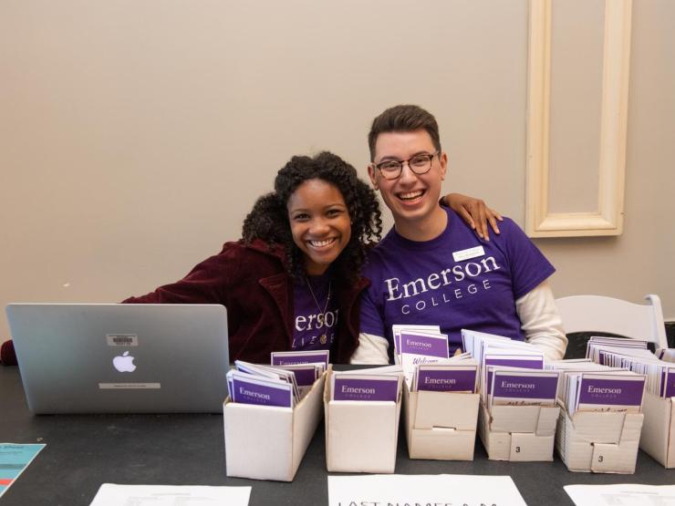 Two students sitting at a table covered in boxes holding welcome leaflets