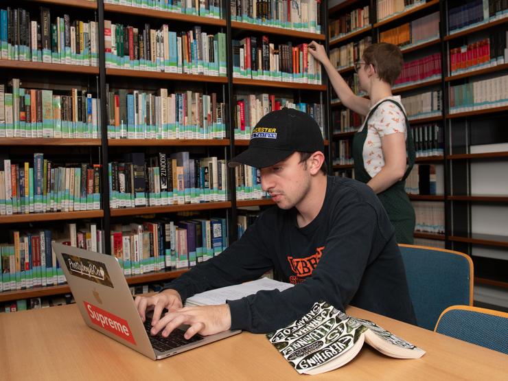 A student works on a laptop in the library while another selects a book