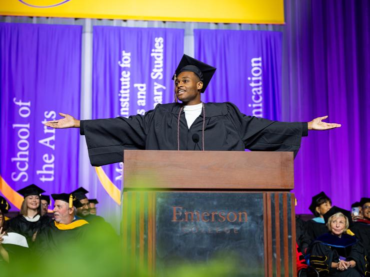 A graduating student at a lectern wearing regalia, arms outstretched at Commencement. Faculty members sit behind them, also dressed in regalia.