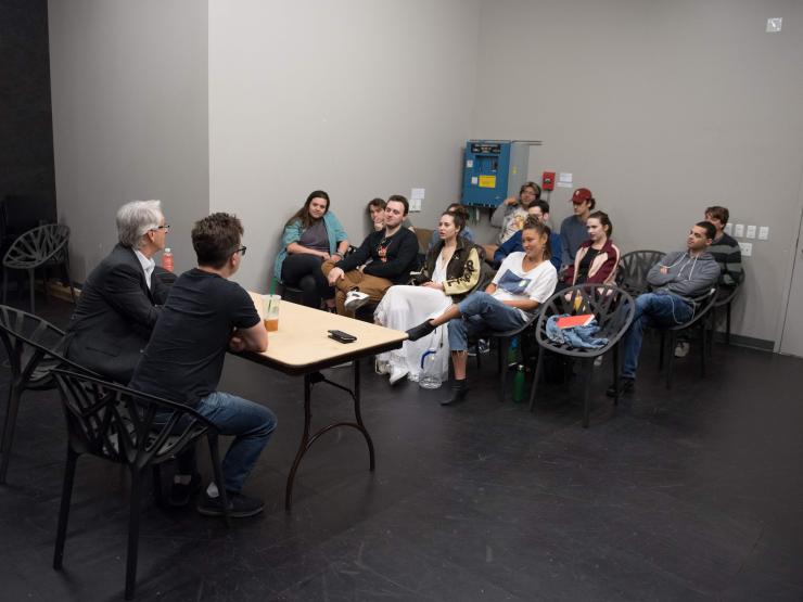 A group of students facing a table in a classroom, with two instructors seated at the front of the table