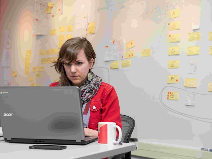A student works intently at a laptop with a mug and phone in front of them, they are seated in front of a large board with many sticky notes