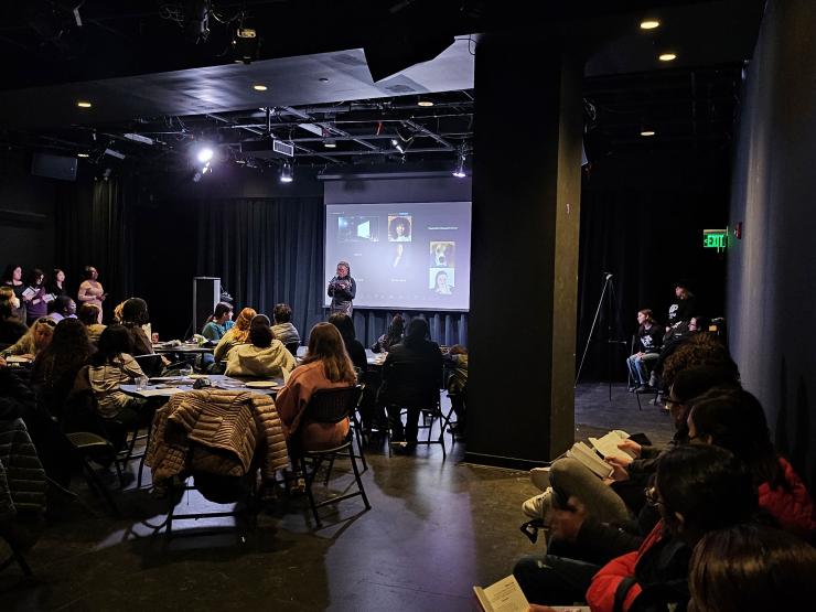 Image of a student reading at a microphone to a room full of people as well as Zoom attendants