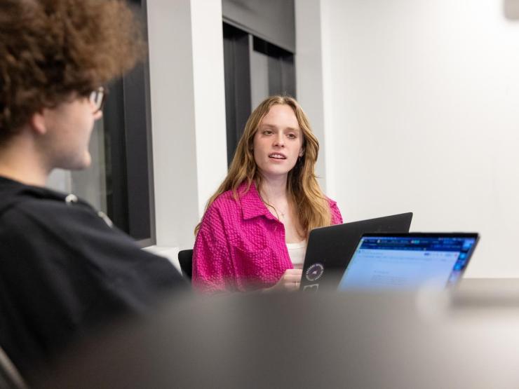 Two students in a classroom, having a discussion while using laptops. One is wearing a pink button-down shirt over a white tanktop, the other is wearing a hoodie.