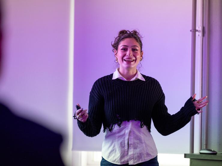 Smiling young woman with curly hair gives a presentation. Wears a white collared shirt under black sweater, standing in front of purple background