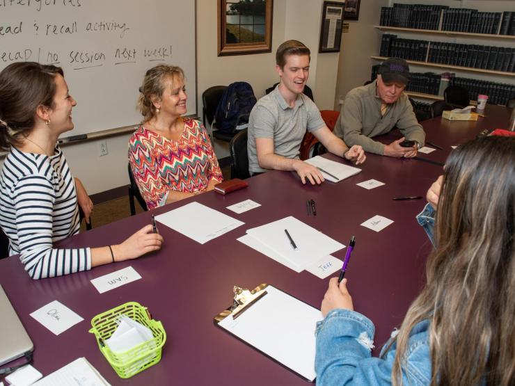 A speech therapy session between a group of people sitting at a table with worksheets. A dry erase board is available behind them.