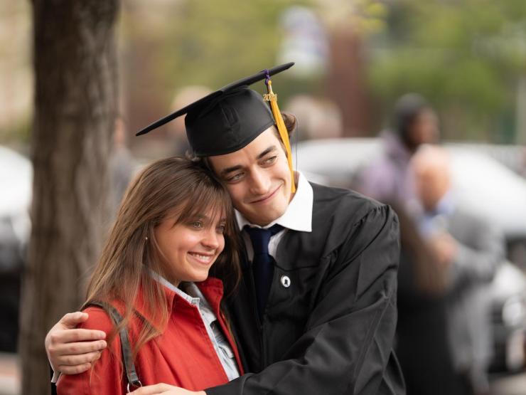 A student embracing a loved one outdoors after their graduation ceremony. They are wearing a cap and gown. Their loved one is wearing a red jacket. They are smiling for a photograph.