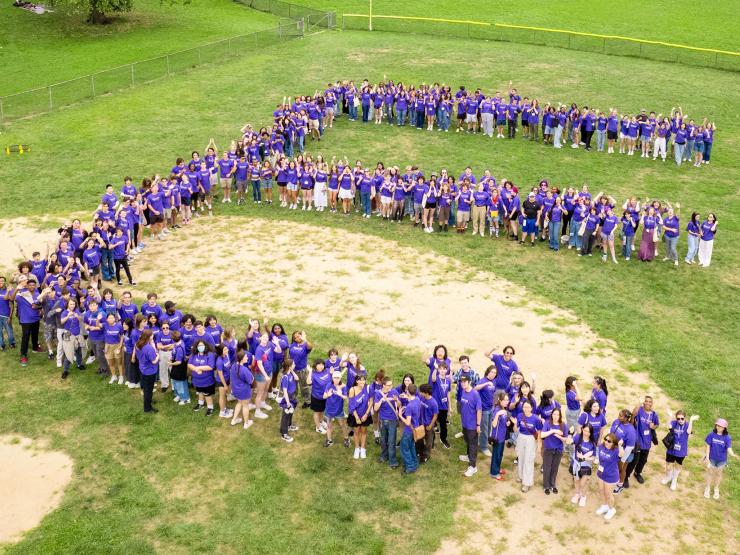 A group of Emerson students outdoors in a baseball field, arranged in a letter 'E". They are wearing purple apparel.