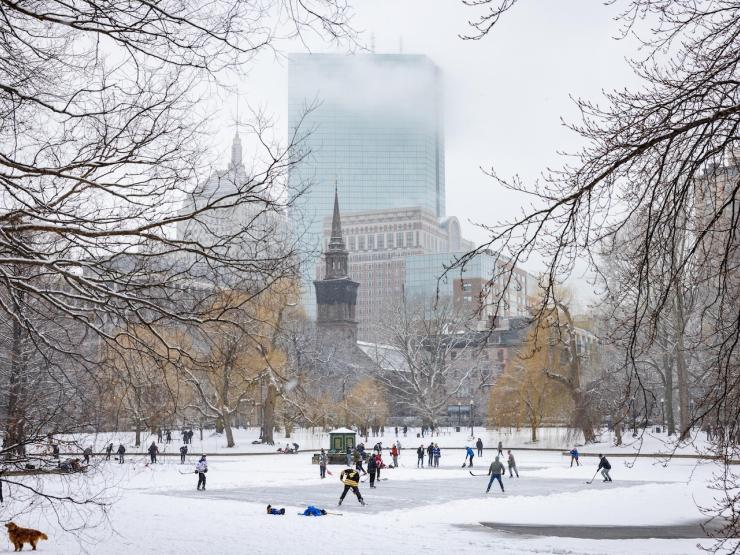 Boston Public Garden in the winter, with people playing hockey on the frozen pond and enjoying the snow, with the Boston skyline in the background.