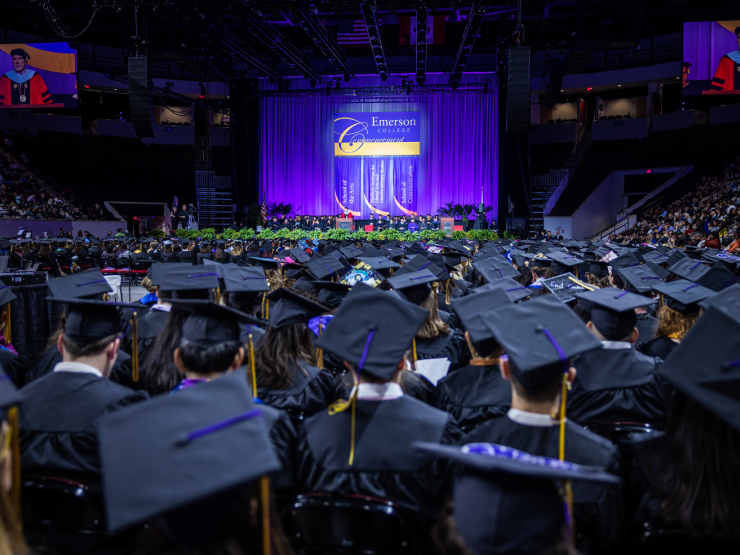 Students in graduation caps and gowns face the stage in a large arena at an Emerson commencement ceremony
