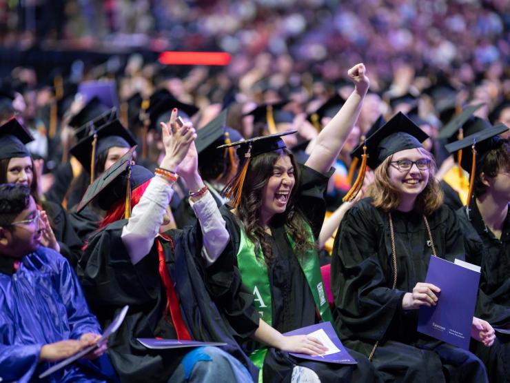Row of students in caps and gowns in a large room of graduates grinning and clapping