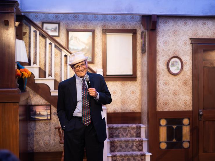 Norman Lear stands onstage, holding a microphone. 