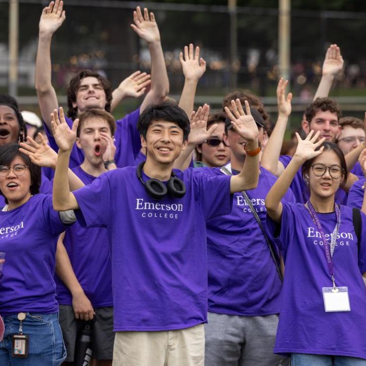 A group of Emerson students stand in a field while waving and cheering, and wearing purple shirts with white text that read Emerson College
