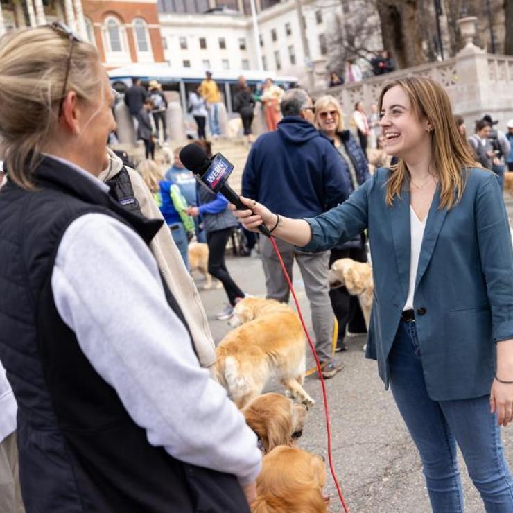 A female student in a blazer interviews a group of people and their golden retriever dogs in downtown Boston.