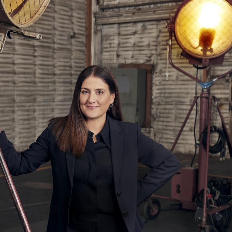 Commencement speaker Pamela Abdy closed mouth smiles with a hand on her hip and another on movie set lighting equipment