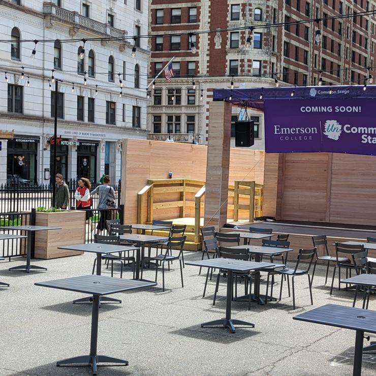 View of the corner of Boylston and Tremont Street, a patio with outdoor tables and chairs are in front of small wooden stage