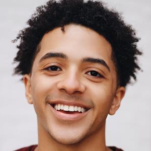A young person with short coily black hair smiles brilliantly in a professional headshot