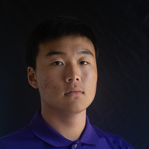 A student looks forward solemnly on a colored background, wearing a purple Emerson polo shirt