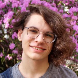 Smiling headshot of a smiling student with glasses and asymmetrical shoulder length hair in front of flowers