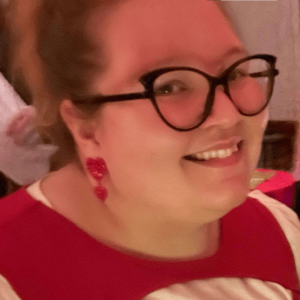 Photo of Mercedes Lamb smiling, her red hair is pulled up and away from her face, on her face she has large black cat eye glasses, and a red dangly heart earing in her ear. She wears a red and while top