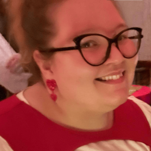 Photo of Mercedes Lamb smiling, her red hair is pulled up and away from her face, on her face she has large black cat eye glasses, and a red dangly heart earing in her ear. She wears a red and while top