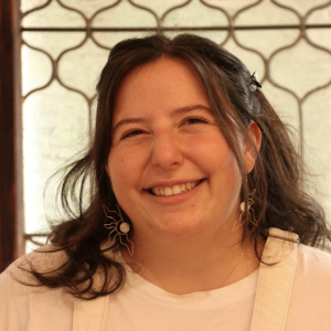 White woman smiling with brown wavy medium length hair, wearing a white t-shirt and white overalls.