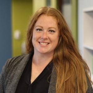 Rebekah Dale sits against a blue and green background wearing a black blouse and grey jacket