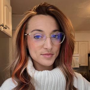 white woman with nose piercing, blonde hair, pink highlights, glasses, white turtleneck