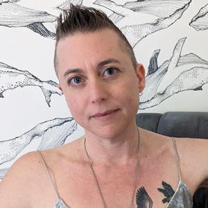 Face and shoulders of Amy L. Clark sitting in front of wallpaper pattered with whales.  Amy is white, small, and in their forties with short hair.