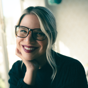 A woman with blonde hair and glasses, wearing a black top, smiles.