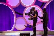 Two students handling a large recording camera on stage during the EVVY Awards, with a colorful purple backdrop behind them