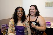 Two people standing next to each other and smiling at an Alumni event. They are standing near a table of swag for attendees. One is carrying a purple baseball cap, branded for Emerson College alumni.