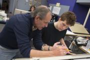 A professor leans over a focused student's drawing desk