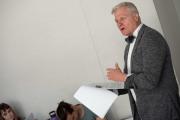 Professor in a blazer and bowtie holds sheets of paper and speaks passionately to a group of students