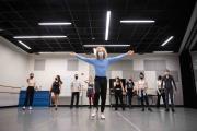 A tap dance instructor wearing a mask in a studio demonstrates with a flourish in front of nine students