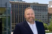 Emerson College President Jay Bernhardt smiles in a blue suit against a backdrop of downtown Boston, specifically the Little and Walker Buildings on Boylston Street