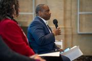 A panelist sits and speaks into a microphone while other panelists watch him.