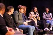 A side view of five panelists on a stage, watching a fellow panelist speak into a microphone.