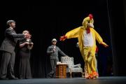 Three actors stand on a stage watching an actor in a chicken costume walking in the foreground.