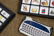 A white wide keyboard and a visual speech aid displaying small pictures of various items, like a red juice box, with basic phrases above each image such as 'I want a drink'