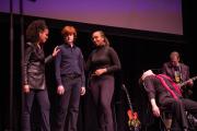 Four students dressed in black clothing act out a scene, a musician plays guitar