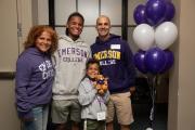 A student with their family smiling at the camera, flanked by purple and white balloons. The group is wearing Emerson College sweatshirts, while a young child holds up a stuffed lion.